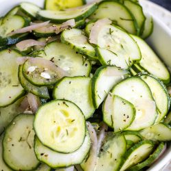 A white bowl filled with sliced cucumbers mixed with thinly sliced onions and fennel seeds, garnished with herbs.