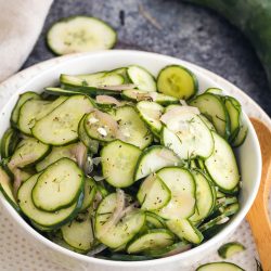 A white bowl filled with cucumber salad, featuring thinly sliced cucumbers and onions, garnished with herbs. Sliced cucumbers are scattered on the surface nearby along with whole cucumbers in the background.