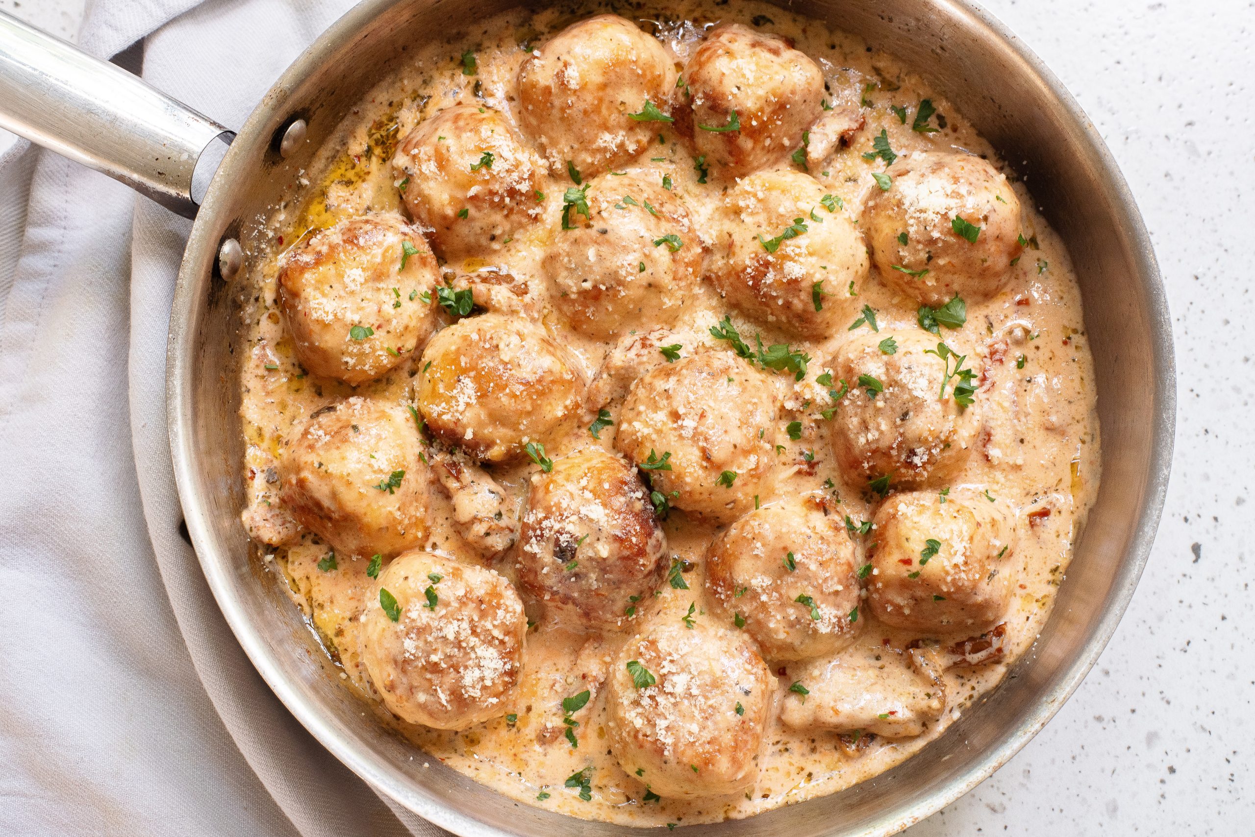 A pan filled with meatballs in a creamy sauce, garnished with chopped parsley and grated cheese.