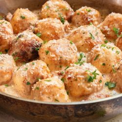 A skillet filled with golden-brown meatballs topped with creamy sauce and garnished with chopped parsley.