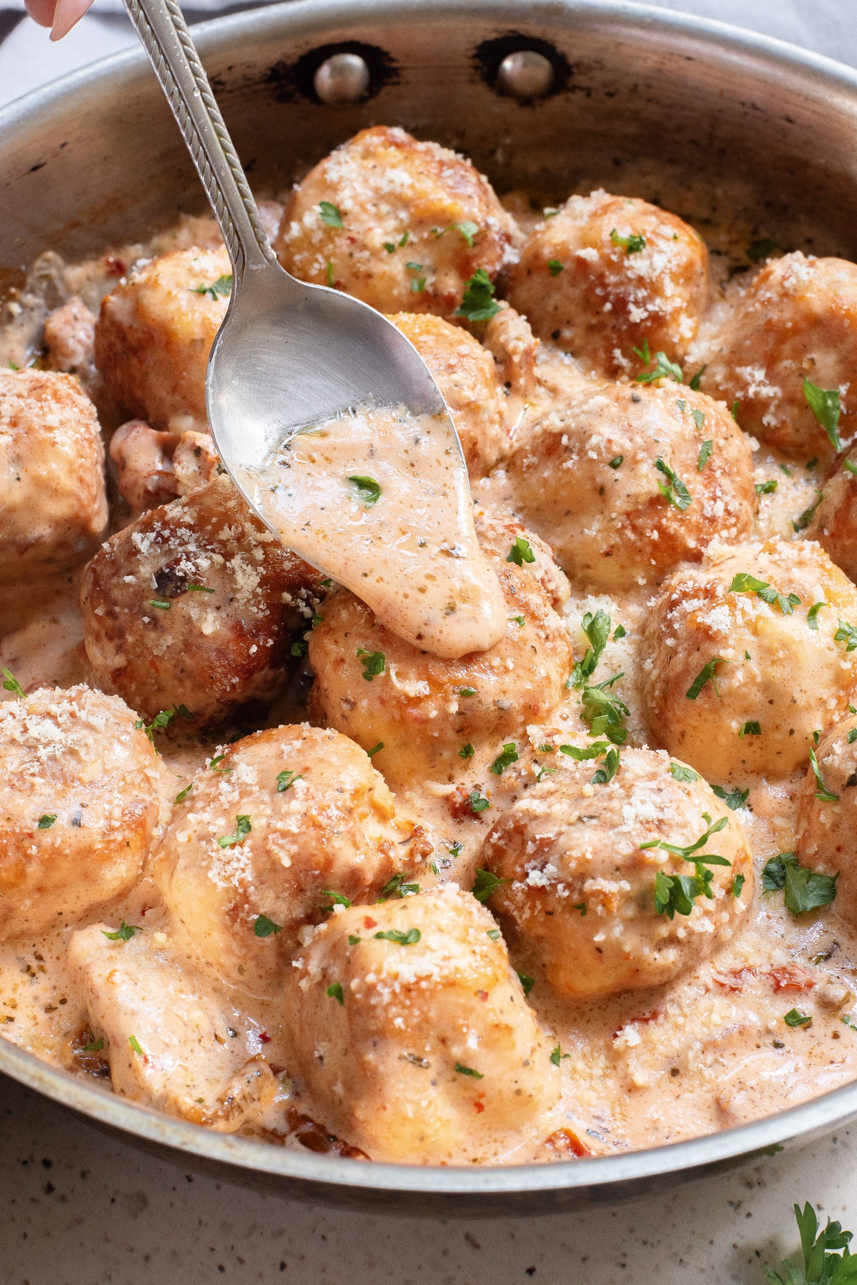 A spoon drizzling creamy sauce over meatballs in a pan, garnished with chopped parsley and grated cheese.