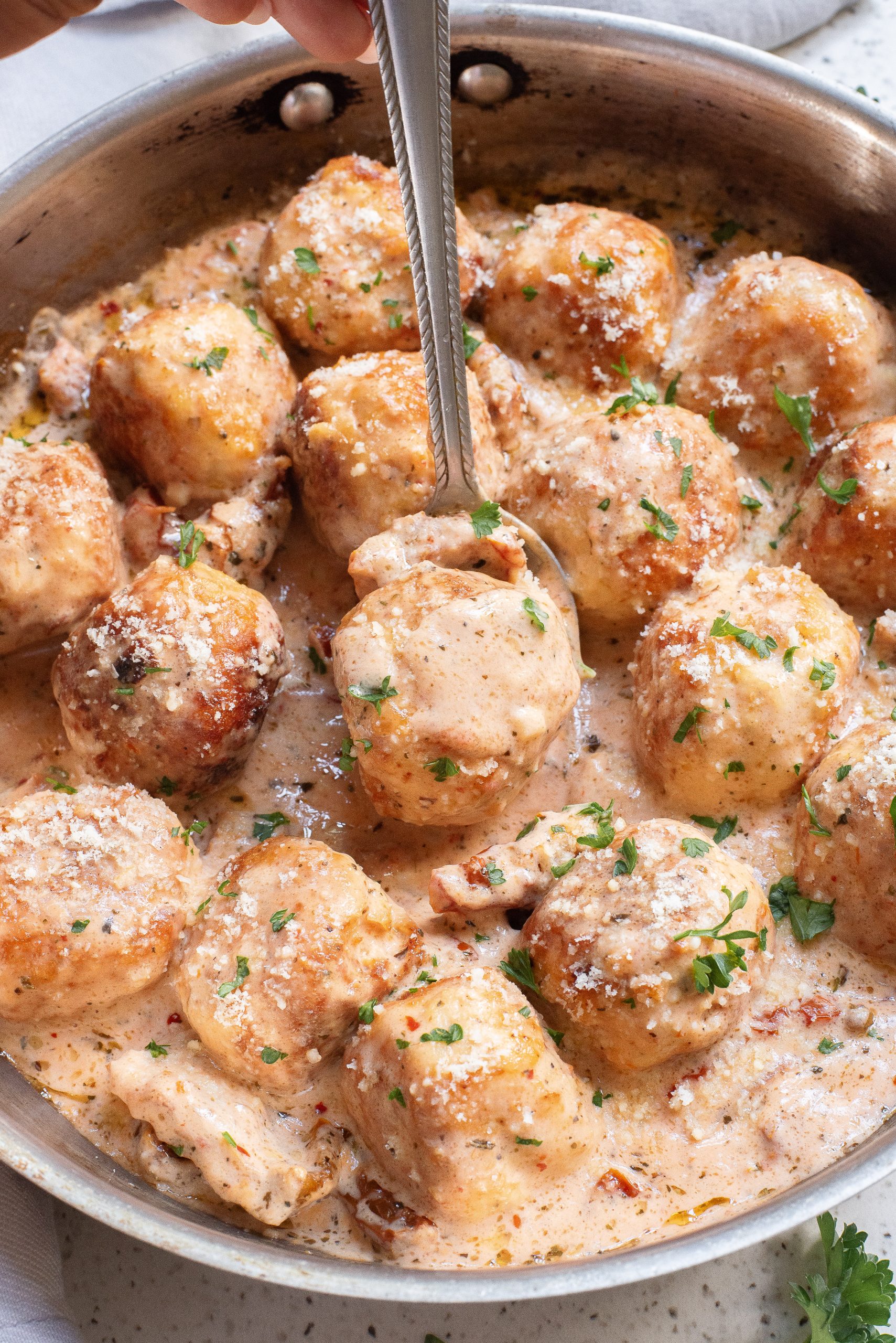 A skillet filled with meatballs in a creamy sauce, garnished with chopped herbs and grated cheese. A spoon is lifting one meatball from the sauce.