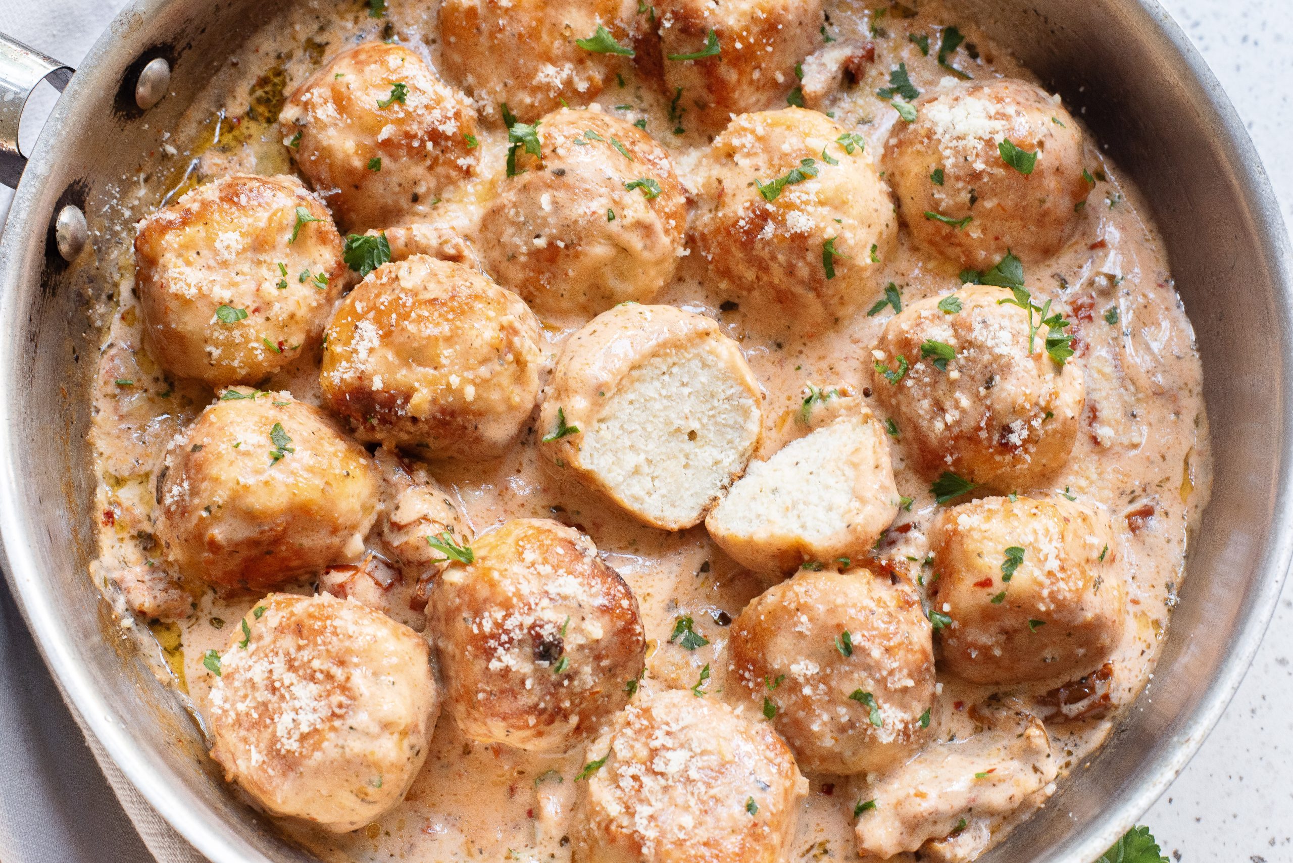 A skillet containing Marry Me Chicken Meatballs in a creamy sauce, garnished with chopped herbs. One meatball is cut in half, revealing the inside.