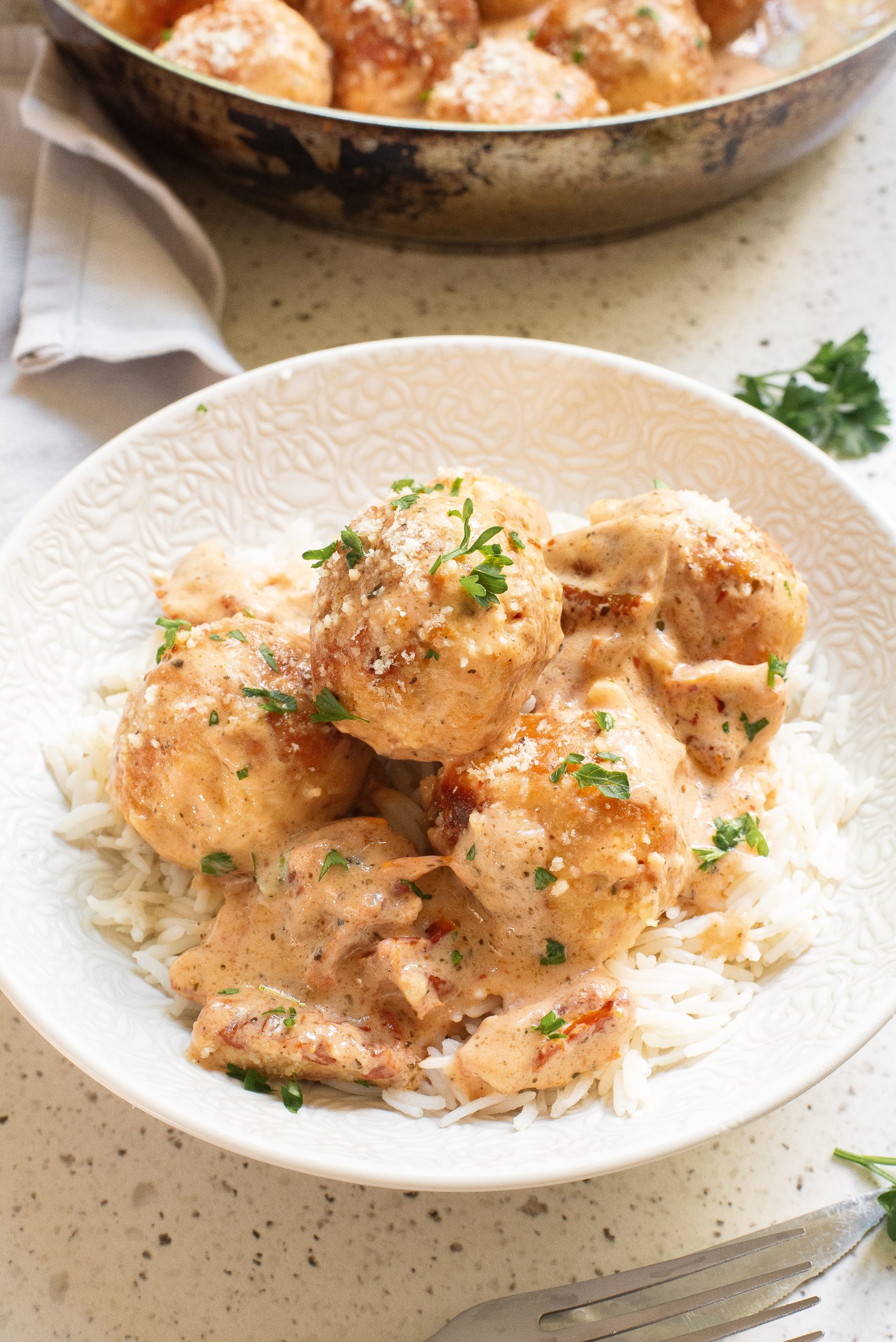 A white bowl containing rice topped with Marry Me Chicken Meatballs covered in a creamy sauce, garnished with chopped parsley, sits on a speckled countertop next to a fork and napkin.