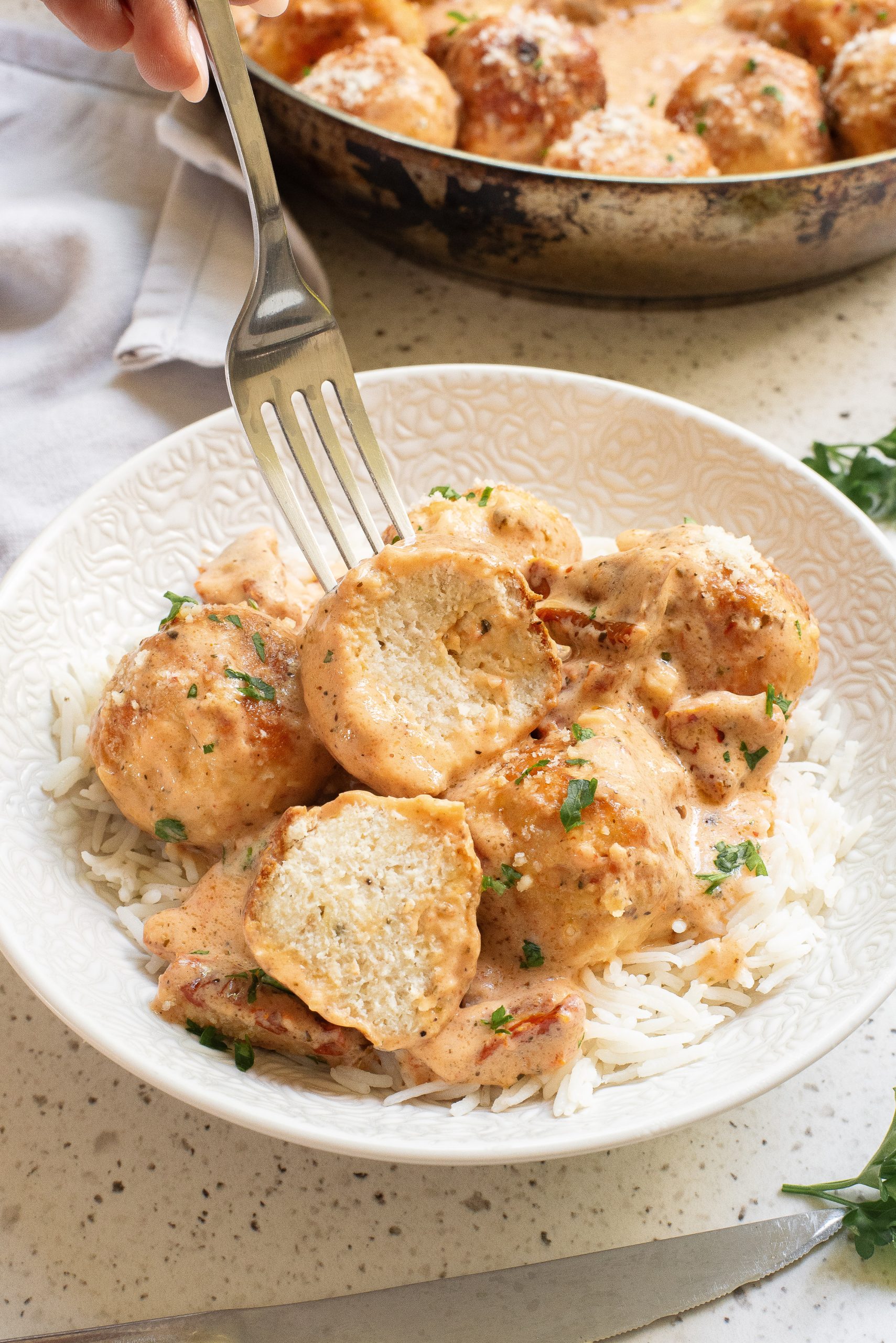 A bowl of rice topped with meatballs in a creamy sauce. A fork is holding a sliced meatball, showcasing its interior. A pan with more sauce-covered meatballs is in the background.