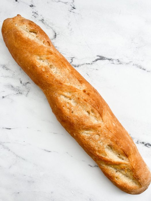 A single loaf of crusty French bread is placed on a marbled surface. The baguette has several diagonal slashes on its golden-brown crust.