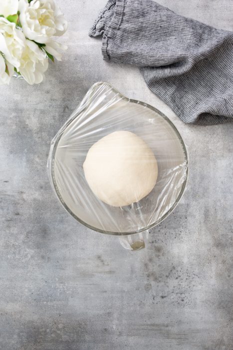 A ball of dough in a glass bowl covered with plastic wrap, placed on a textured gray surface. A gray cloth and white flowers are in the background.