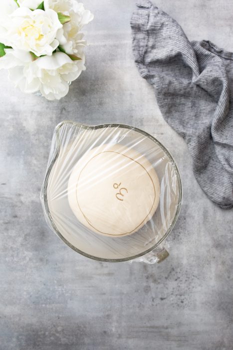 A bowl with dough covered in plastic wrap on a grey surface. A white flower bouquet and a striped cloth are in the background.