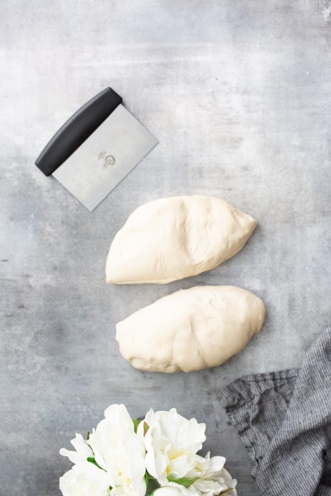 Two pieces of dough are placed on a textured grey surface alongside a metal dough scraper and a grey-striped cloth. White flowers are partially visible in the bottom corner.