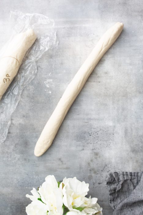 A roll of dough is placed on a grey surface next to white flowers and a piece of crumpled plastic wrap.