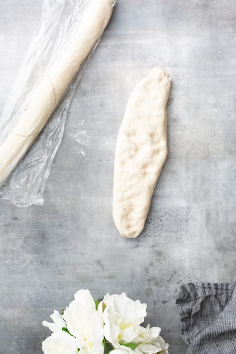 A rolled piece of dough beside a plastic-wrapped roll of dough on a gray surface. White flowers and part of a gray cloth are visible in the lower portion of the image.