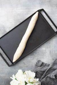 An elongated piece of dough lies on a black baking tray, with a gray fabric and white flowers nearby.