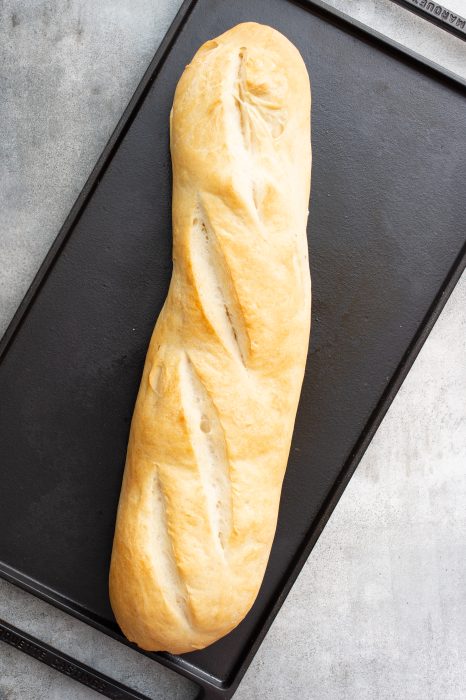 A freshly baked loaf of French bread with a golden crust and three diagonal slashes, placed on a black baking tray.