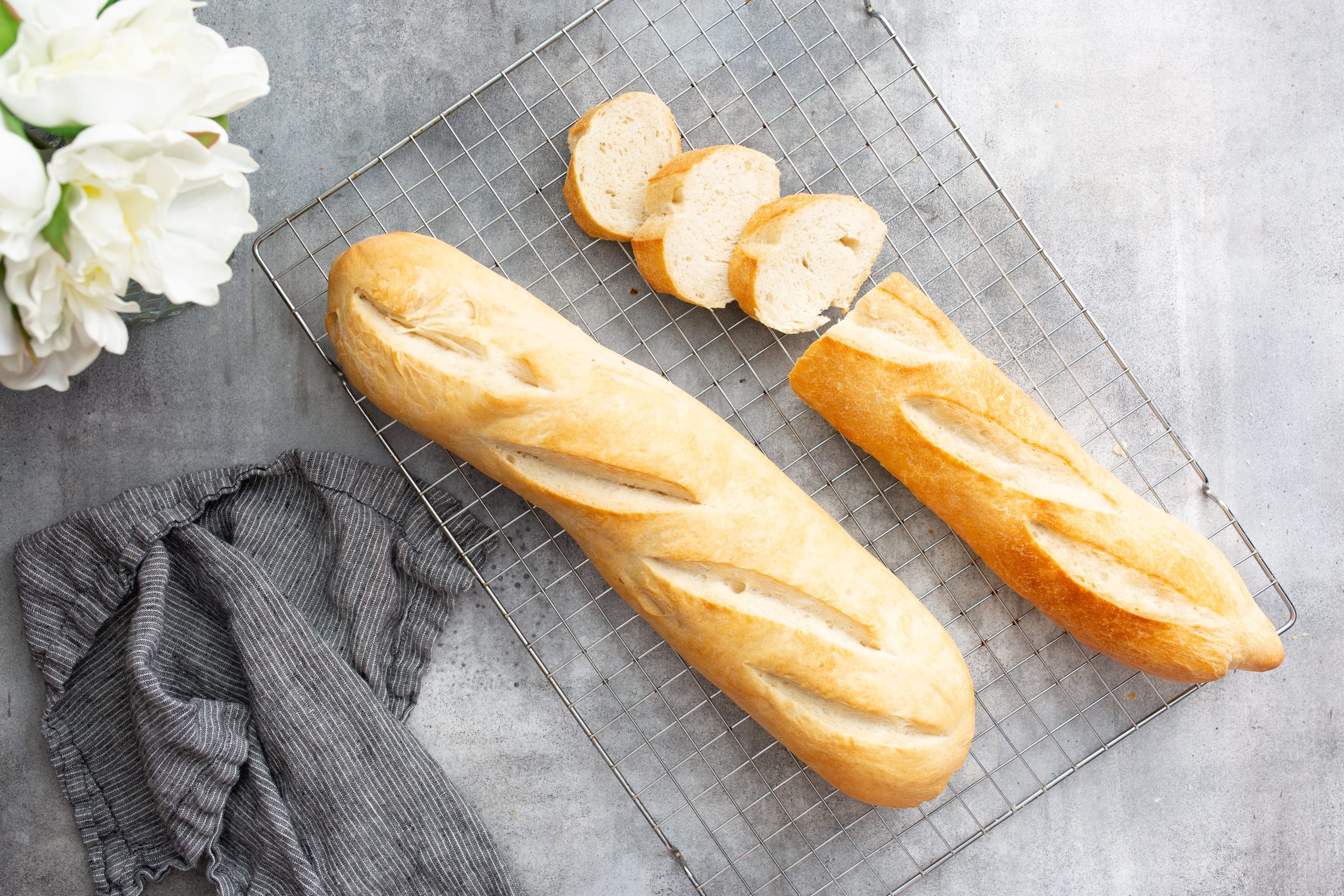 Two freshly baked baguettes, one partially sliced, on a cooling rack. A gray cloth and white flowers are placed nearby on a gray surface.