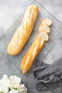 Two freshly baked baguettes, one whole and one partially sliced, rest on a cooling rack. A dark gray cloth and a white flower are placed nearby on a gray surface.