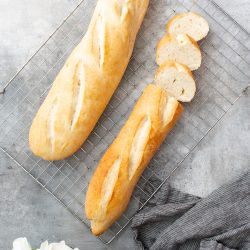 Two freshly baked baguettes, one whole and one partially sliced, rest on a cooling rack. A dark gray cloth and a white flower are placed nearby on a gray surface.