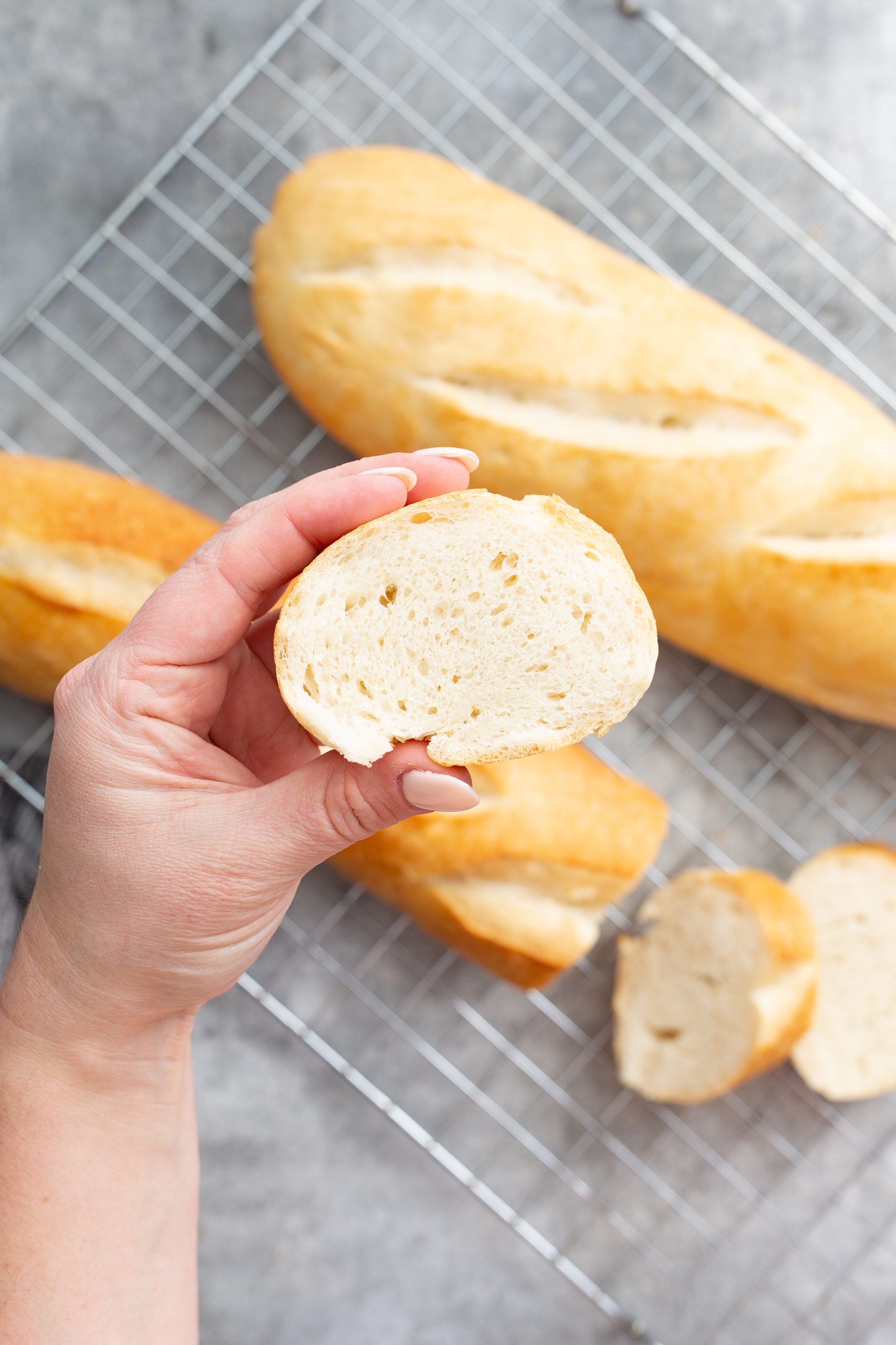 A hand holding a slice of bread in front of several loaves and slices of bread on a wire cooling rack.