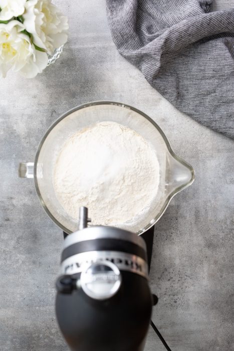 A mixing bowl filled with flour on a countertop near a stand mixer, a gray fabric, and a vase of white flowers.