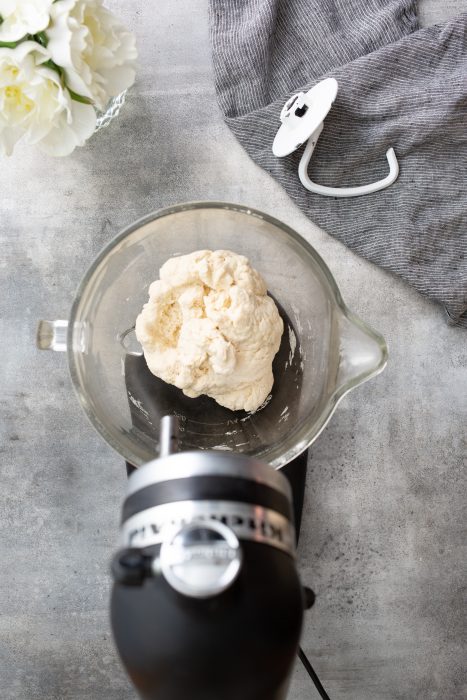 A stand mixer with dough in a glass bowl, a dough hook attachment on a gray cloth, and a vase with white flowers are on a gray countertop.