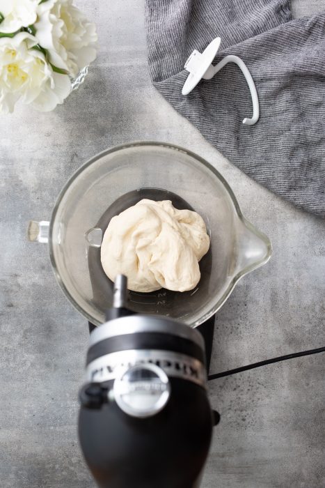 Dough in a mixing bowl attached to a stand mixer. A gray cloth and a white floral arrangement are nearby on a concrete surface.