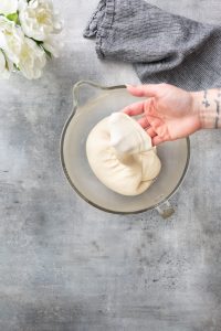 A hand stretches dough in a glass bowl on a gray surface, with a blue cloth and white flowers visible in the background.