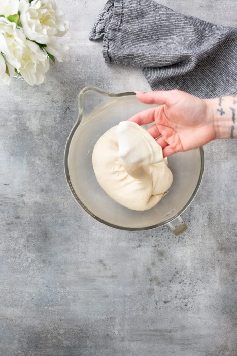 A hand stretches dough in a glass bowl on a gray surface, with a blue cloth and white flowers visible in the background.