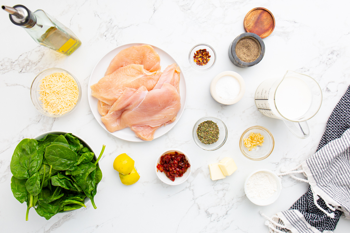 A flat lay of various cooking ingredients on a white surface, including raw chicken, spinach, spices, cheese, butter, garlic, flour, lemon, sun-dried tomatoes, milk, and oil.