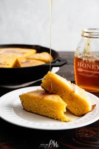 A slice of cornbread is served on a white plate with honey drizzling over it. A jar of honey and more cornbread in a skillet are in the background.