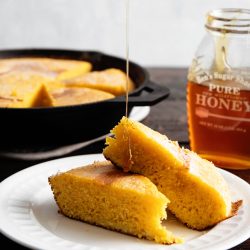 A slice of cornbread is served on a white plate with honey drizzling over it. A jar of honey and more cornbread in a skillet are in the background.