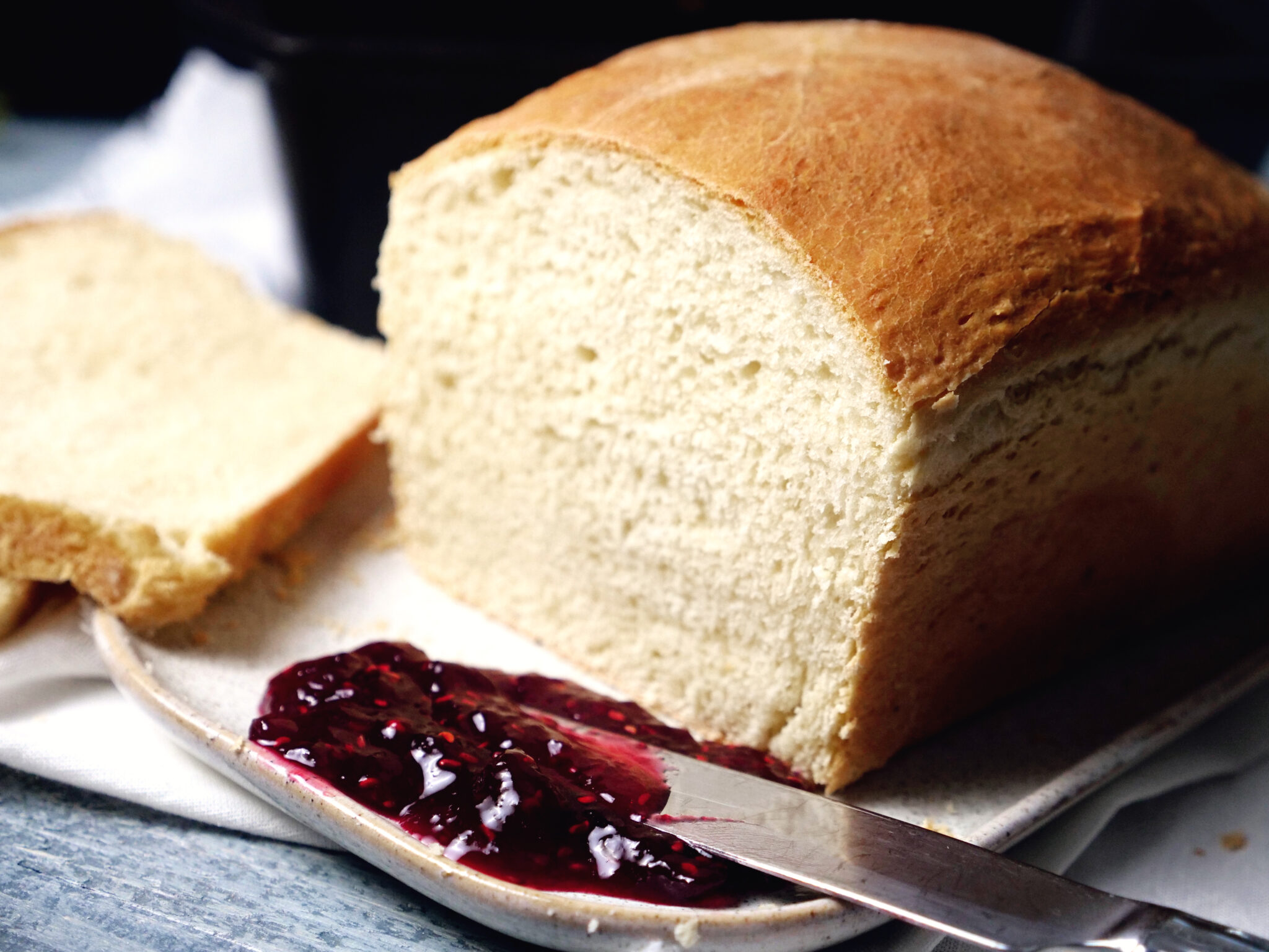 A loaf of homemade bread sits on a plate with a knife and some berry jam. One slice of bread has been cut.