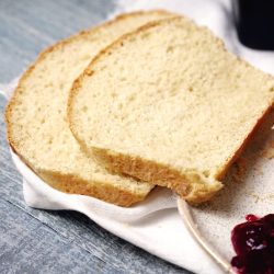 Two slices of white bread are placed on a white cloth next to a dish of red jam on a blue wooden surface.