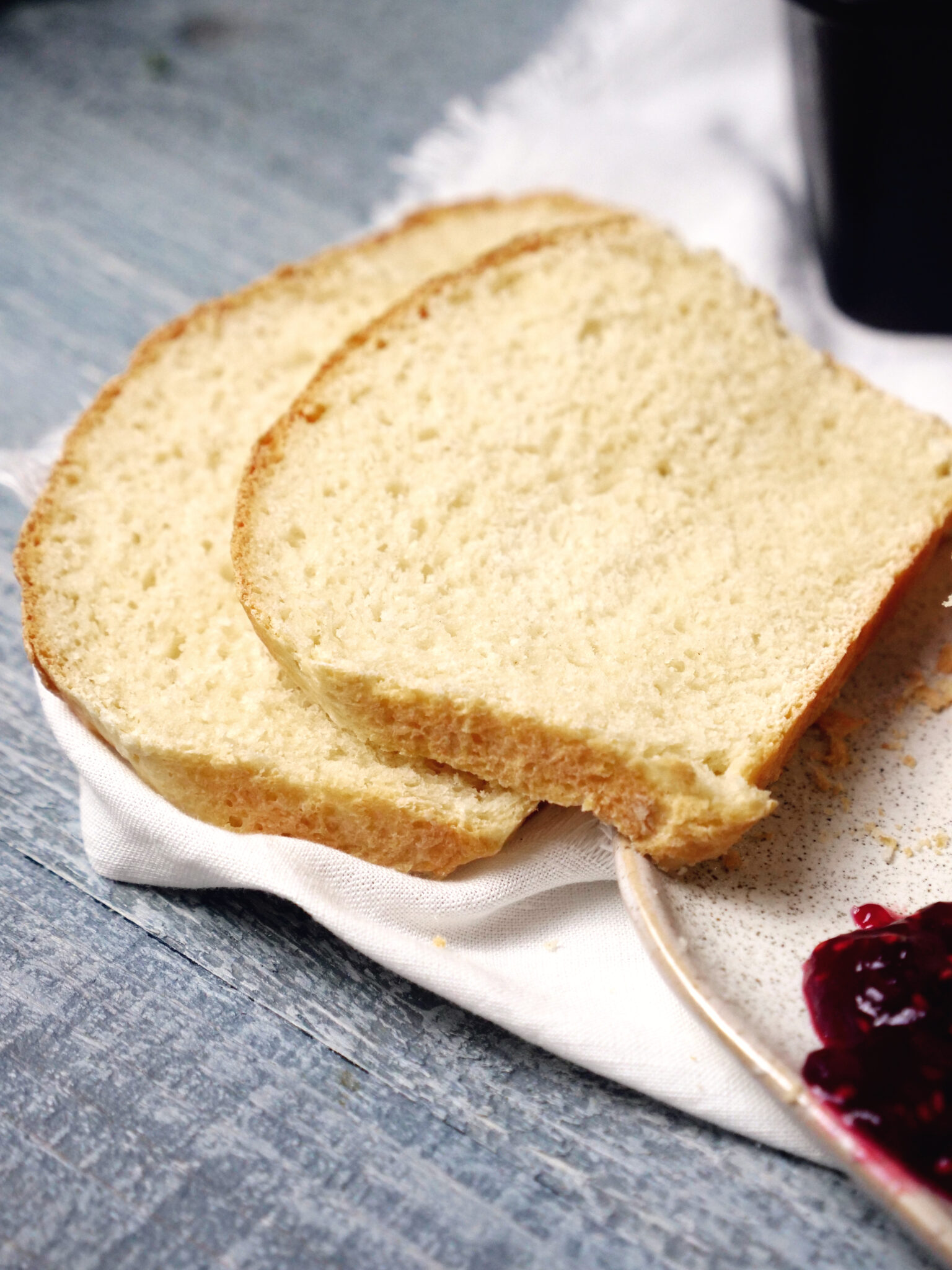 Two slices  are placed on a white cloth next to a dish of red jam on a blue wooden surface.