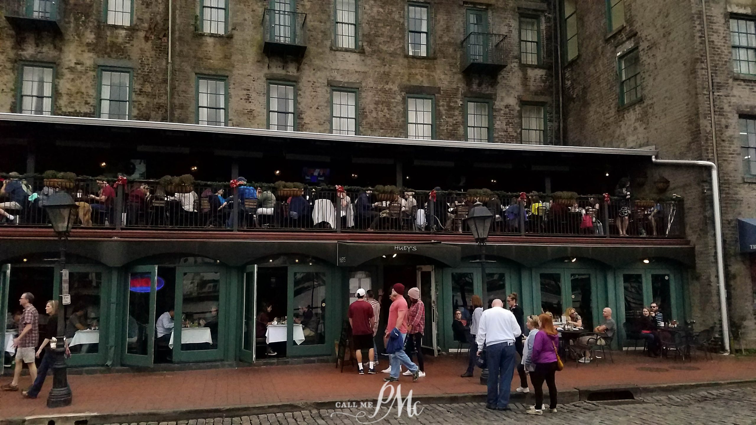 People are gathered outside and on the balcony of a rustic building housing a café. Several green double doors and windows are visible, with pedestrians walking on the cobblestone street below.