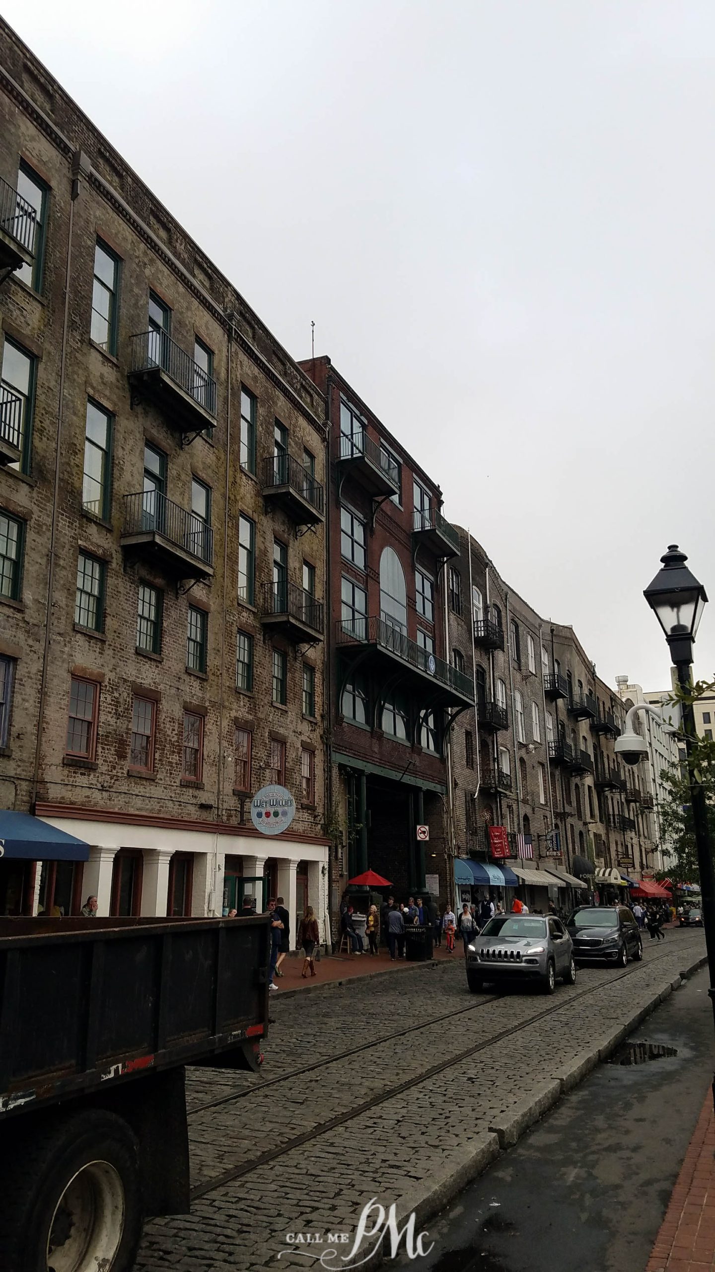 48 hours in Savannah. A cobblestone street lined with historic brick buildings, some with balconies. People are walking along the sidewalk, and cars are parked on the street. A streetlamp is visible in the foreground.