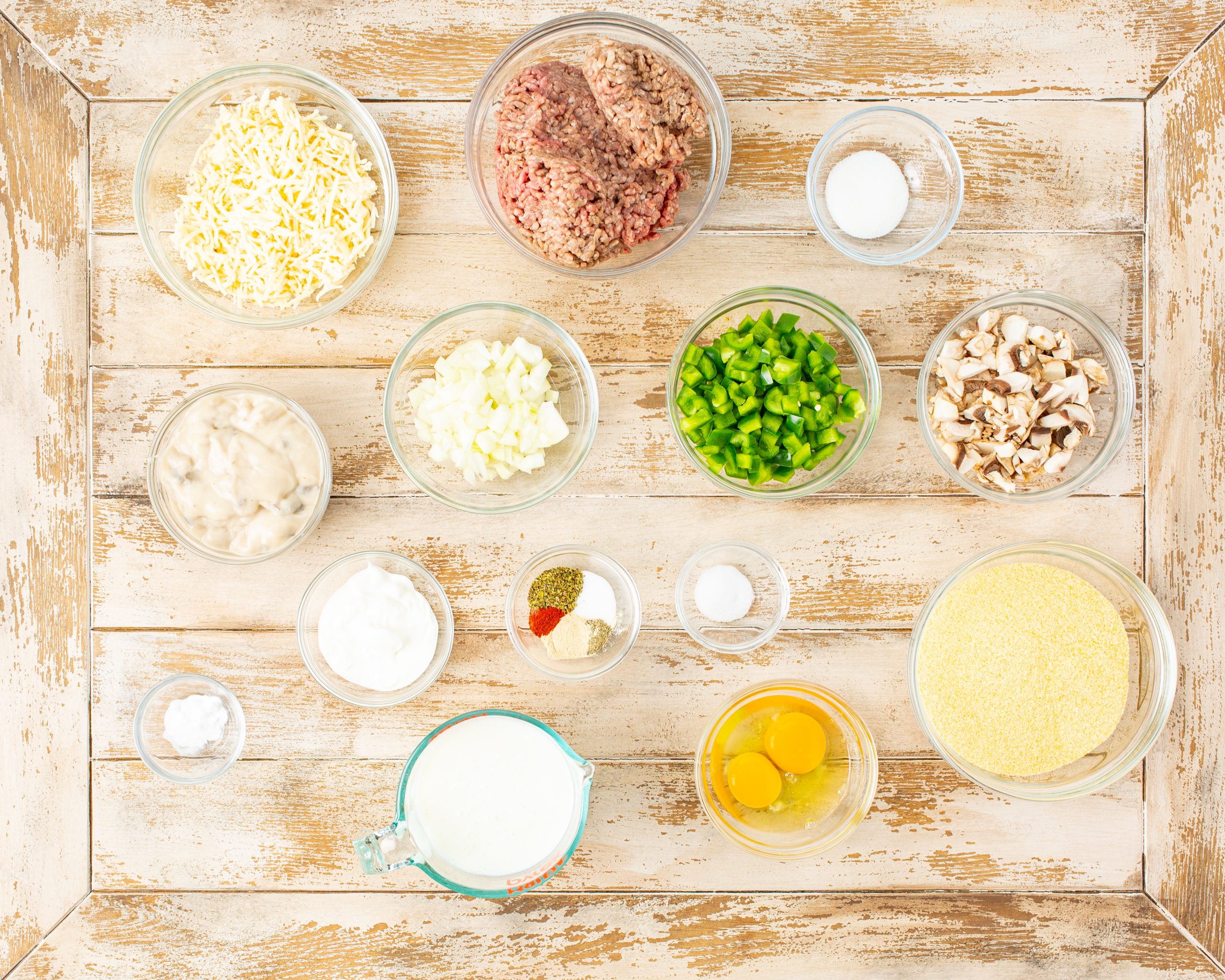 Ingredients for cooking on a wooden surface, including shredded cheese, ground meat, salt, chopped green peppers, mushrooms, onions, eggs, cream of mushroom soup, spices, and milk.