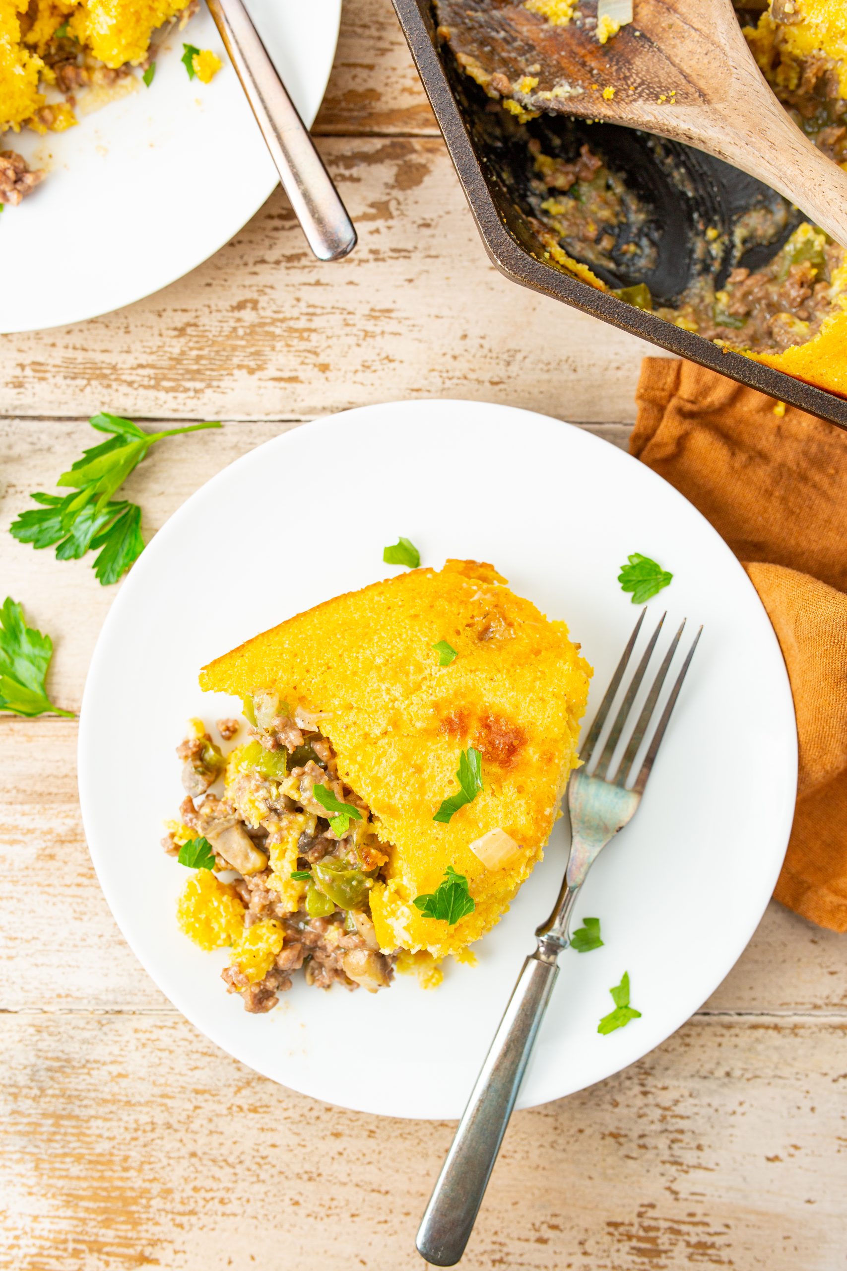 cornbread casserole with ground beef topped casserole with meat and vegetables on a white plate with a fork, garnished with parsley, and next to a baking dish with a wooden spoon.