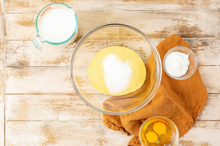 Ingredients for a batter are laid out on a wooden surface. They include a bowl of dry mix, a measuring cup with milk, two eggs in a small bowl, and a ramekin with a white substance.