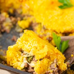 Close-up of a casserole dish containing a portion of cornmeal pudding mixed with beef and vegetables, topped with garnished parsley leaves. A wooden spoon is partially visible in the dish.