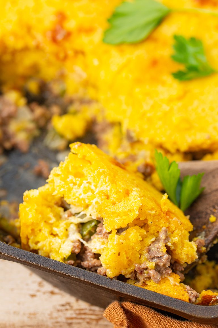 Close-up of a casserole dish containing a portion of cornmeal pudding mixed with beef and vegetables, topped with garnished parsley leaves. A wooden spoon is partially visible in the dish.