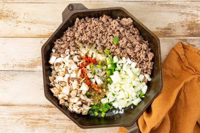 A cast iron skillet on a wooden surface contains cooked ground meat, chopped mushrooms, diced onions, and various spices. An orange cloth is placed beside the skillet.