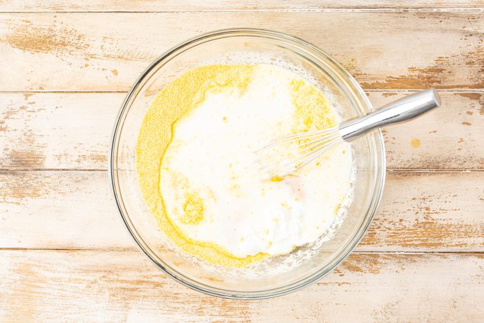 A glass bowl of yellow dry ingredients and liquid being mixed with a metal whisk on a wooden surface.