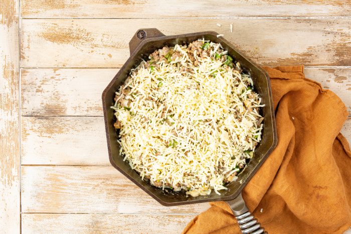 A cast iron skillet filled with a mixture of ground meat and vegetables, topped with shredded cheese, is placed on a wooden surface next to an orange napkin.