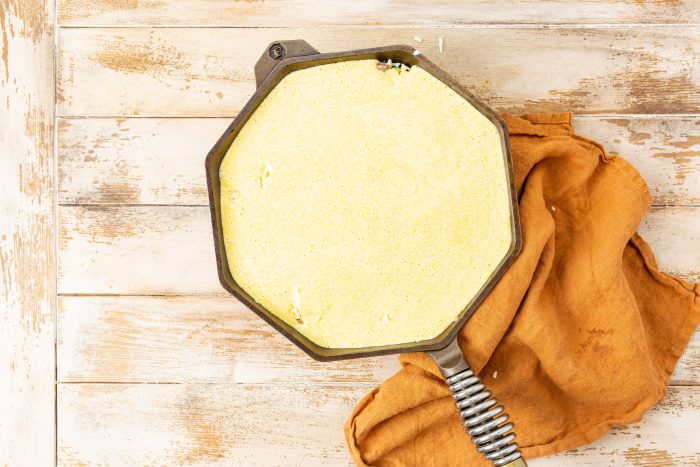 A round, yellow cornbread batter in a cast iron skillet rests on a rustic wooden surface, with an orange cloth partially underneath.