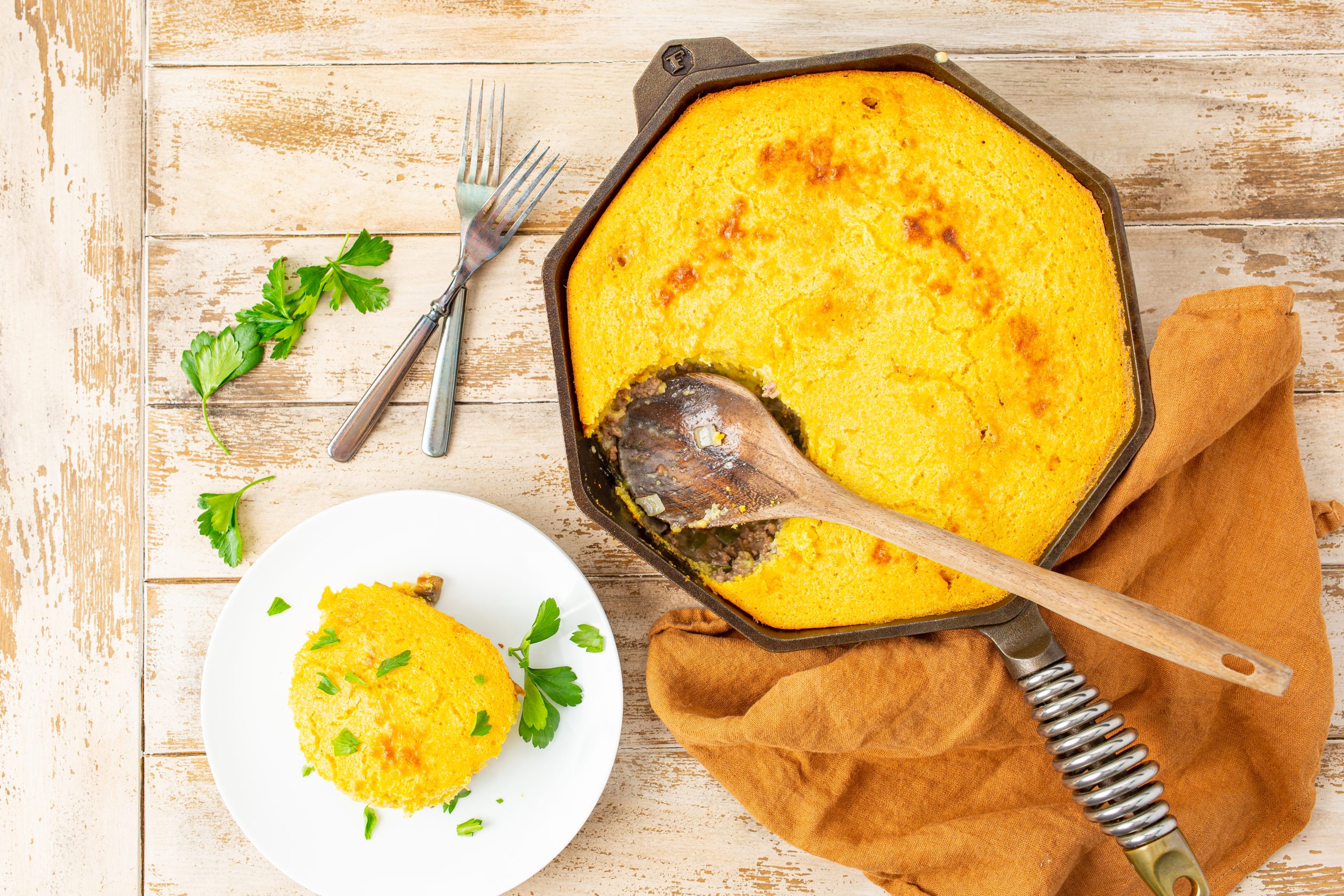 An overhead view of cornbread casserole with ground beef with a serving spoon, a slice served on a white plate with parsley garnish, and two forks on a wooden table.