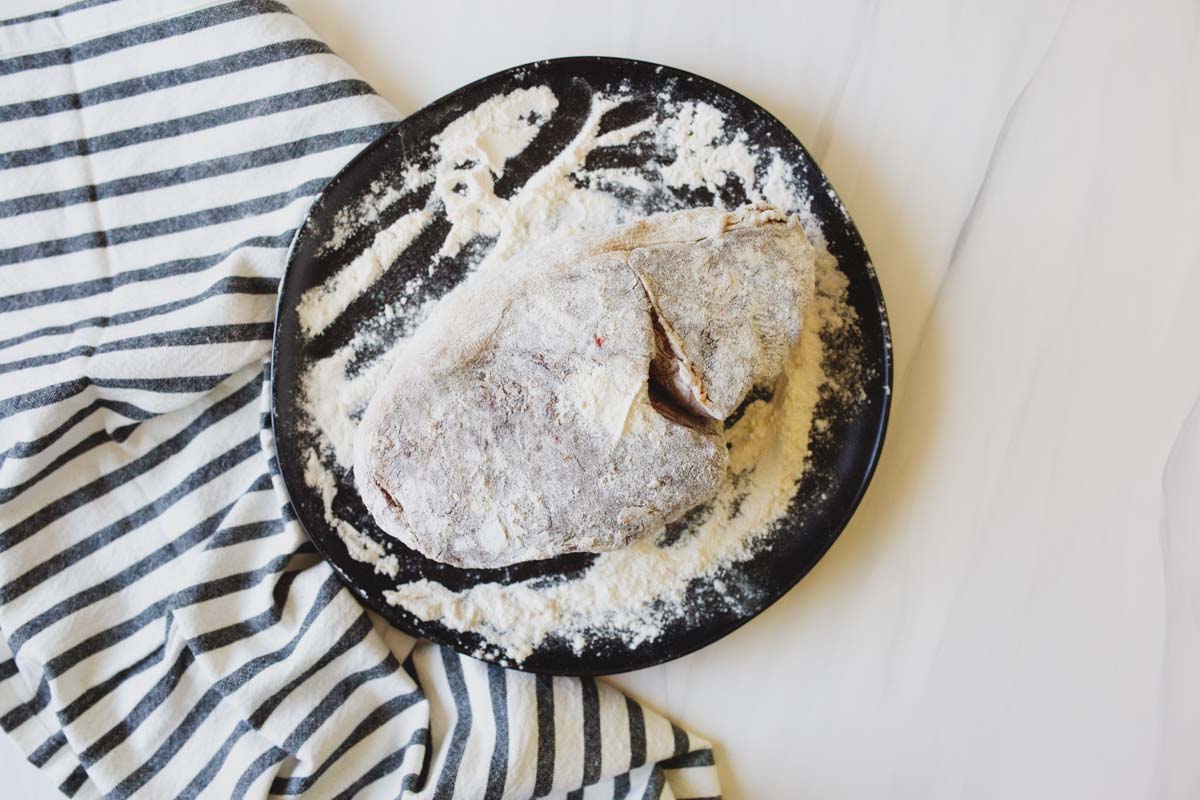 A rustic loaf of bread covered in flour rests on a black plate beside a striped cloth on a light-colored surface.