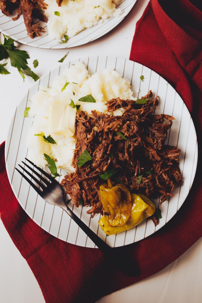 A plate with shredded Dutch Oven Mississippi Pot Roast, mashed potatoes topped with butter, and pickled peppers is placed on a red cloth, with a black fork beside it.