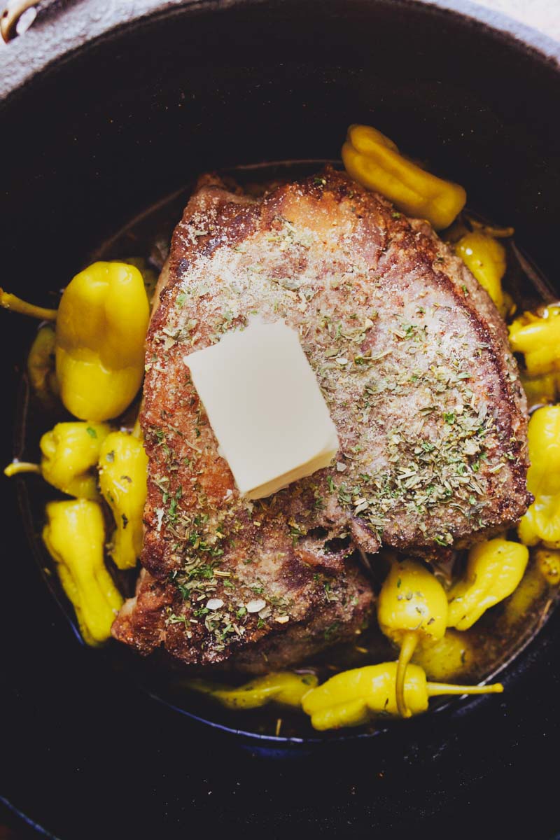 A cooked piece of meat topped with a pat of butter and sprinkled with herbs, surrounded by yellow peppers, in a cast iron skillet.