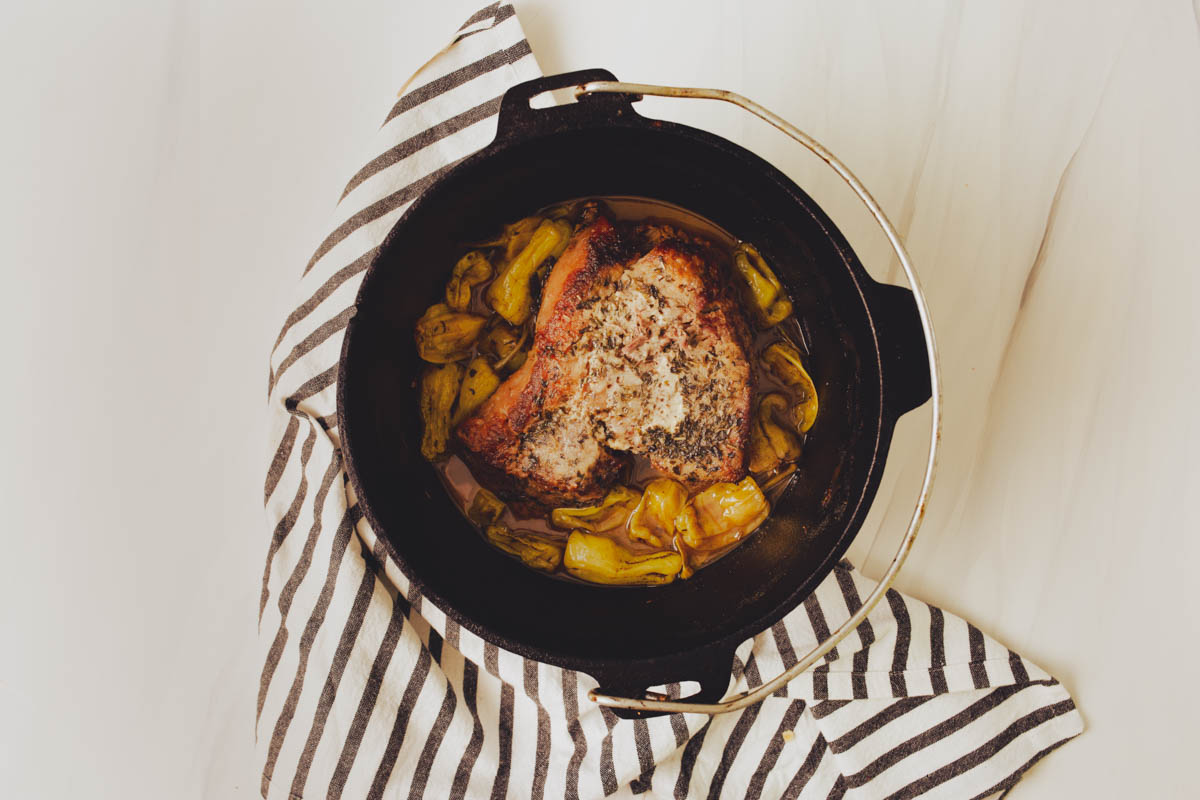 A cooked seasoned Dutch Oven Mississippi Pot Roast, surrounded by vegetables, sits in a black iron pot on a striped cloth.