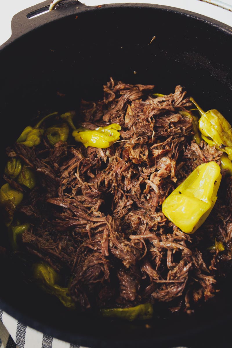 Close-up of shredded beef with whole yellow peppers in a large black pot.