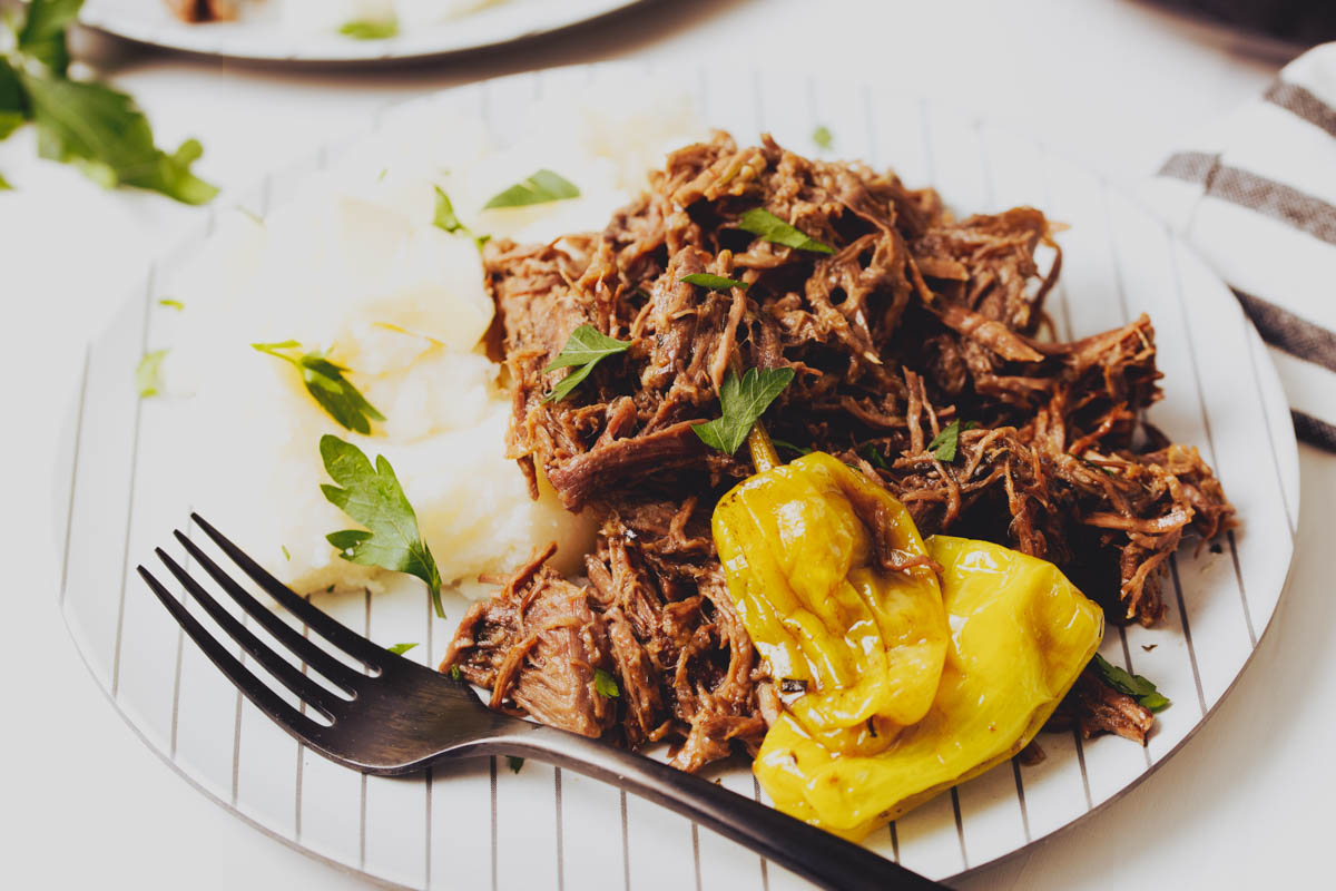 A plate of shredded beef, mashed potatoes, and pepperoncini garnished with parsley, with a black fork resting on the plate.